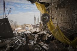 Rubble and debris are strewn about in a destroyed operating room in a hospital with a yellow tarp dangling from the ceiling alongside a hanging light lamp. The outer wall of the operating room is missing, exposing the operating room to the outside.