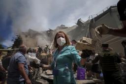 A nurse in blue scrubs wearing a medical face mask is standing in the foreground of a destroyed hospital surrounded by other civilians and military personnel. Smoke is billowing from behind near a building with the roof caved in.