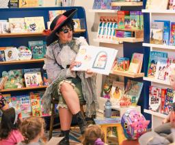 A striking, heavily made-up individual dressed in a black hat and long ruffled jacket as if like a storybook pirate sits amid shelves of children’s books reading to young children.