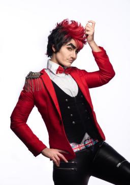 A man in an extravagant red jacket with fancy epaulets, a black vest and black leather pants affects a saucy pose and expression. His dark hair has a shocking red swoop on top and his heavy, pasted-on eyebrows are glittery.