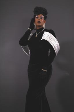 A glamourous Black woman poses with a gloved hand to her face. She is wearing a black and white velvet dress, glitzy diamond jewelry and dramatic makeup.