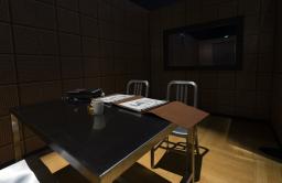 A foreboding, dimly lit space contains two metal chairs and a metal table on which a file and a coffee cup sit.