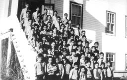 Dozens of young Indigenous students are dressed in a boys’ uniform of a shirt, tie and slacks in this archival black and white photo. They stand on the stairs leading into a school building.