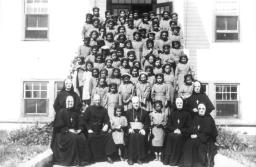 Dozens of young Indigenous students are dressed in a girls’ uniform of a dress and tam in this archival black and white photo. They stand on the stairs leading into a school building. At the forefront of the group are five nuns wearing habits and two priests in hassocks.