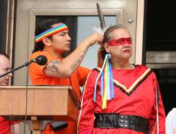 Behind a podium and microphone, an Indigenous man with a rainbow headband affixes a feather to the hair of a person dressed in red with a painted red band across their eyes.