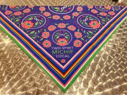 A purple piece of fabric is adorned with pale red and blue flowers and Métis symbols. The border is green, white, pink, blue, yellow and orange stripes. The words “Two-Spirit Michif Local” are embroidered in a corner.