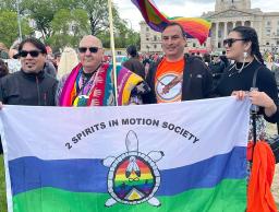 Four Indigenous people at a demonstration hold a flag that depicts a turtle, at the centre of which are Two-Spirit symbols, including two eagle feathers joined together, against a rainbow background. A rainbow flag flutters behind the four people.