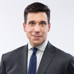A white man with dark hair smiles slightly into the camera. He is wearing a dark suit with an off-white shirt and a dark blue tie with small white polka dots.