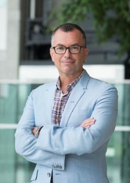 A white man with short hair and eyeglasses smiles towards the camera. He is wearing a light blue-grey blazer and multicoloured dress shirt.