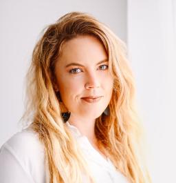 A white woman with long blond hair and a nose ring looks into the camera. She is wearing a white shirt.