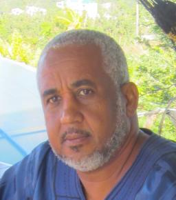 A Black man with greying black hair and beard looks into the camera with greenery behind him. He is wearing a blue shirt.