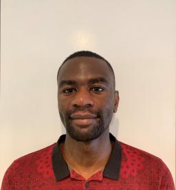 A Black person with very short black hair looks into the camera with a slight smile. They are wearing a red shirt with a printed black line drawing design and a black collar.
