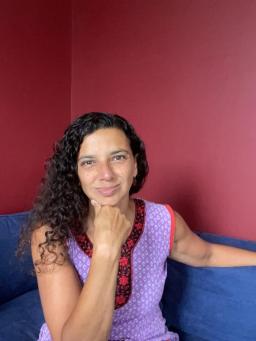 A person with long black curly hair smiles at the camera. Her chin is resting on her right hand and she is sitting on a blue sofa. The wall behind her is dark red.