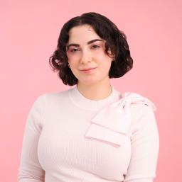 A white woman with brown hair smiles towards the camera. She is wearing a light pink sweater with a pink bow over her left shoulder.