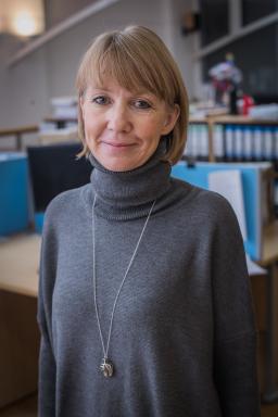 A white woman with blonde hair smiles towards the camera. She is wearing a long gray turtleneck sweater and a silver necklace and pendant.