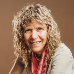 A white-appearing woman with curly blond hair smiles at the camera. She is wearing a beige shirt and a red patterned scarf.