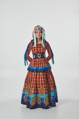 A full-length studio portrait of a person standing against a plain light background, wearing a vibrant Indigenous regalia red patterned dress with blue trim and a wide black belt decorated with large circular silver accents. She is also wearing a feathered headdress with blue and white details. Her hands are clasped behind her back, and she is facing forward.