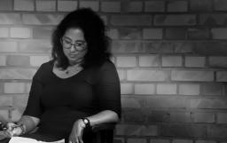 A black-and-white portrait of a woman with dark curly hair who is looking down at a paper on her lap.