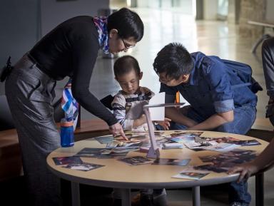 A woman and man bend over a table where a young child is playing with a pile of photographs.