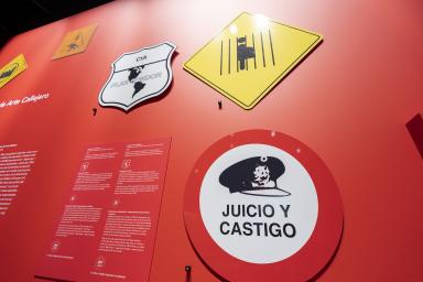 Signs with graphic designs and Spanish text hang on a red wall. The larges sign says “Juicio Y Castigo.”