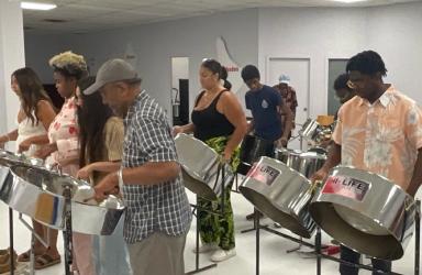 Nine people stand in rows playing steel drums.