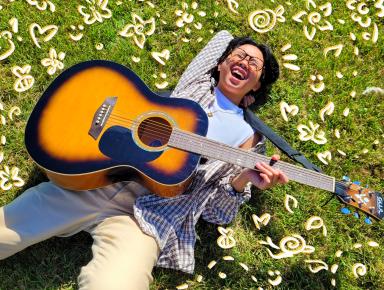 Kris Cahatol lies laughing on green grass and is surrounded by computer-drawn flowers and hearts. Kris is holding a guitar across their chest.