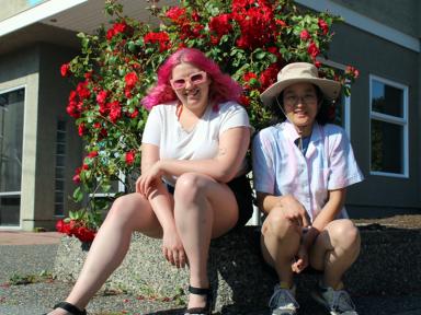 Tiana Garcia and Megumi Kimata sit smiling in front of a red rose bush. Garcia has long fuchsia hair and pink sunglasses. Kimata is dark-haired and wears round glasses and a hat.