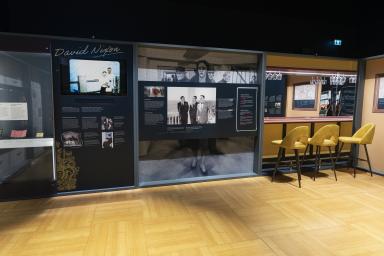 A museum display depicts three distinct sections. Two have photos and text and the third is a mock-up of a bar with a counter and three yellow chairs.