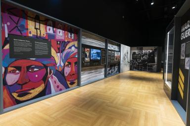 A museum gallery, in which the most visible panel is a colourful art piece depicting Indigenous faces and symbols.