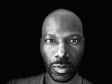 A black-and-white close-up portrait of a Black man looking straight at the camera with a neutral expression.