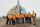 Ten people in hard hats and safety vests stand in front of a large, unique-looking building. The grounds are muddy and unfinished.