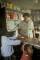 A woman stands behind a wooden store counter. Two young children are standing in front of her. One faces the woman behind the counter while the other looks away.