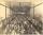 A black and white photo of a movie theatre audience. The picture is taken from the front of the theatre looking towards the back, so the faces of the audience can be seen. Potted palm trees line the walls on each side.