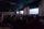 A seated audience viewing three large screens in a dimly lit room.