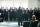 A choir of men wearing black outfits hold books and sing in front of a conductor.