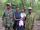 A woman is holding a baby, while four men in army fatigues stand beside and behind her. They are all standing in front of a forest, posed for the camera.