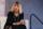 A woman sitting on a chair with hands clasped looks thoughtfully ahead, as if answering a question.