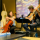 A cello player and guitar player chat and play music together in a café.