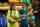 Two guitar players in bright and colourful outfits sing into microphones in front of a balloon arch.