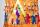 A section of an orange jingle dress is shown. Jingles, or small silver cone-shaped pendants, are visible at the top of the image. Under them, seven human figures are shown, all in various colours and sizes. One of the figures is wearing an orange shirt. These human figures are holding hands inside a multicoloured tipi.