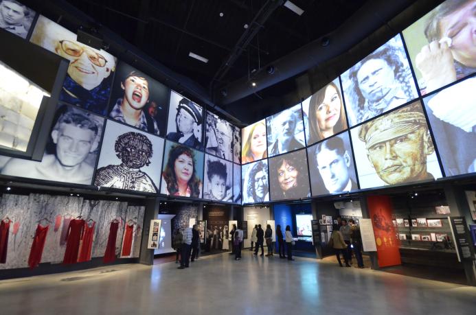 People explore a Museum gallery with two rows of square portraits above a series of alcoves, containing photographs, videos, and text. Partially obscured.