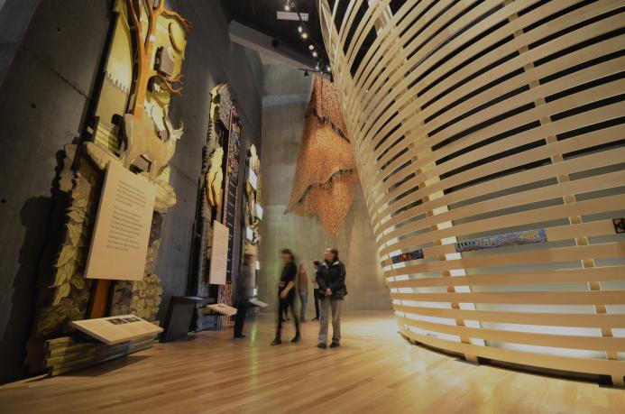 People in a Museum gallery explore tall wooden panels with wildlife and Métis beadwork. There are curved horizontal wooden slats to the right.