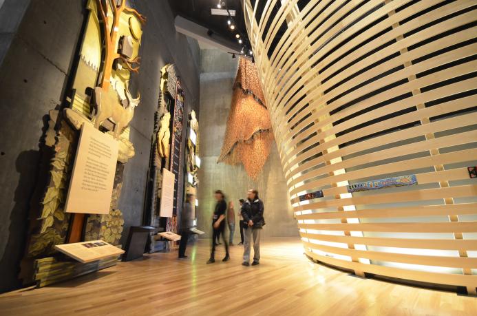 People in a museum gallery explore tall wooden panels with wildlife and Métis beadwork. There are curved horizontal wooden slats to the right. Partially obscured.