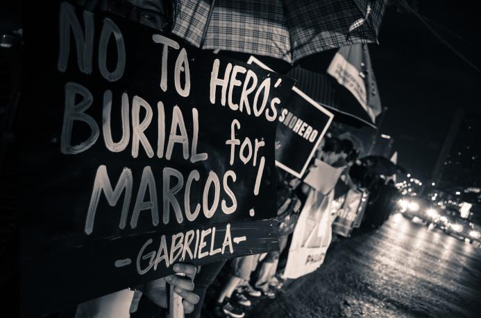 People standing in the rain under umbrellas along a roadside. Headlights from vehicles passing them are on the right side of the image. In the left foreground a sign blocks their faces from view and reads “No to hero’s burial for Marcos! Gabriela.” Partially obscured.