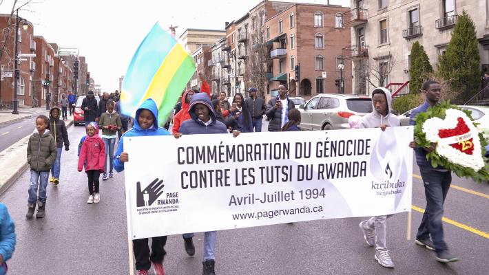 A group of people walking down a street with sign that reads “Commémoration du génocide contre les Tutsi du Rwanda. Avril-Juillet 1994. www.pagerwanda.ca”. Partially obscured.