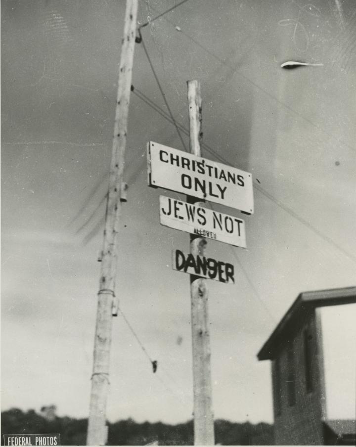 Black and white photo of three signs on a post, reading “Christians only,” “Jews not allowed” and “Danger.” Partially obscured.