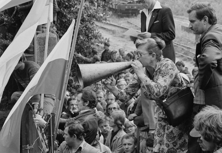 A woman wearing a dress cups her hand to a megaphone placed at her mouth. She is standing elevated above a crowd of people at a gate, with two flags waving nearby. Partially obscured.