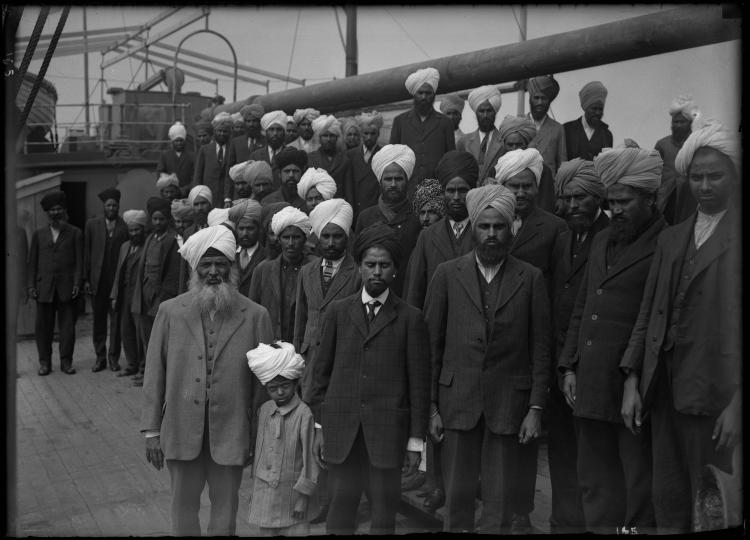 A black and white image of a large group of Sikh men and one young boy standing on the deck of a ship. They are all facing the camera and are not smiling.