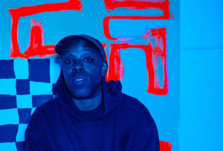 A blue-toned image of a black man wearing a cap and dark hoodie in front of a checkered backdrop with thick red lines. Partially obscured.