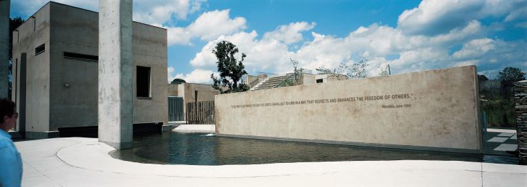 A view of the Apartheid Museum. In the foreground is a pool of water. Rising out of the pool is a large wall with a quote from Nelson Mandela written on it. It reads: “To be free is not merely to cast off one’s chains, but to live in a way that respects and enhances the freedom of others.”